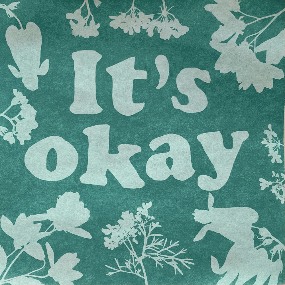 A cyanotype print with the words It's okay in the middle and shapes of flowers around the edge